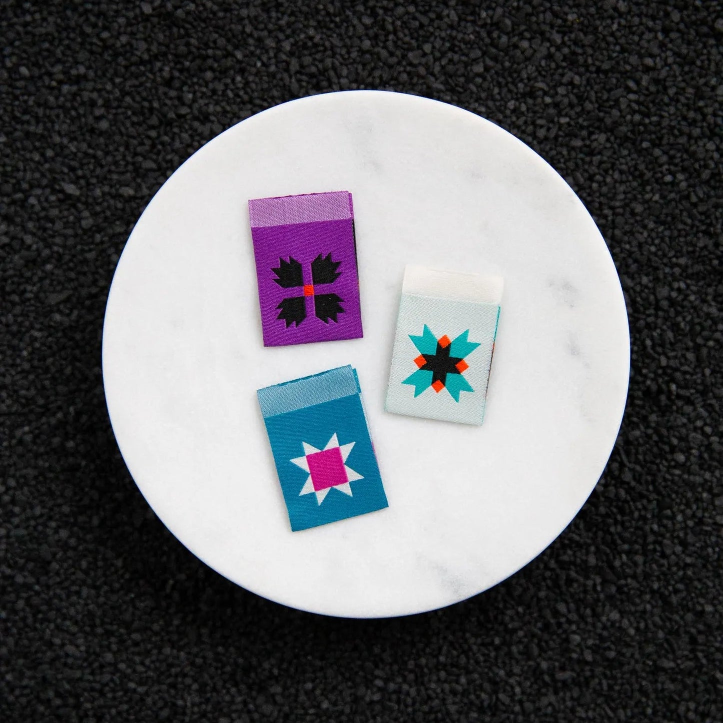 Three small colorful rectangular woven labels arranged on a round white marble plate against a black textured background. The set includes a purple piece with black floral motifs, a light blue piece with a pink starburst, and a white piece with a red and teal starburst.